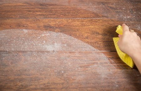 Um Kratzer zu vermeiden sollte Parkett mit einem weichem Baumwolltuch abgewischt werden. Das Bild zeigt einen staubigen Holzfußboden, von dem eine Hand mit Lappen den Schmutz entfernt