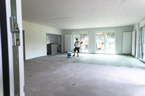 Eine Person grundiert mit einer Malerrolle den Boden eines großen, unmöblierten Raums mit weißen Wänden und bodentiefen Fenstern; in der Nähe ist ein großer Eimer mit der Grundierung zu sehen.