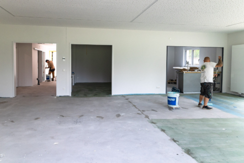 Zwei Arbeiter renovieren ein unfertiges Zimmer; einer streicht eine Wand in der Nähe eines Fensters, der andere arbeitet in einem angrenzenden Raum.