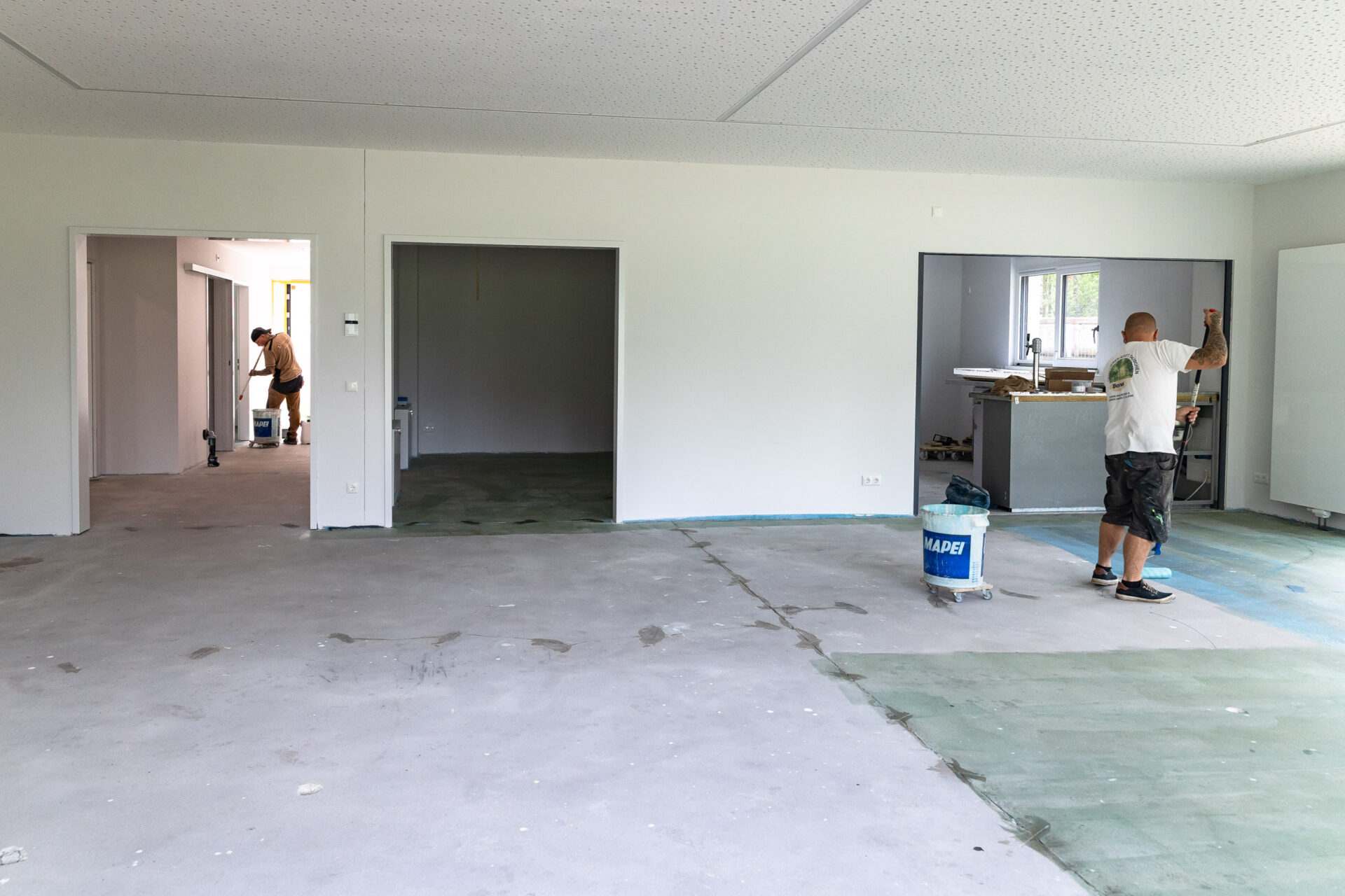 Zwei Arbeiter renovieren ein unfertiges Zimmer; einer streicht eine Wand in der Nähe eines Fensters, der andere arbeitet in einem angrenzenden Raum.