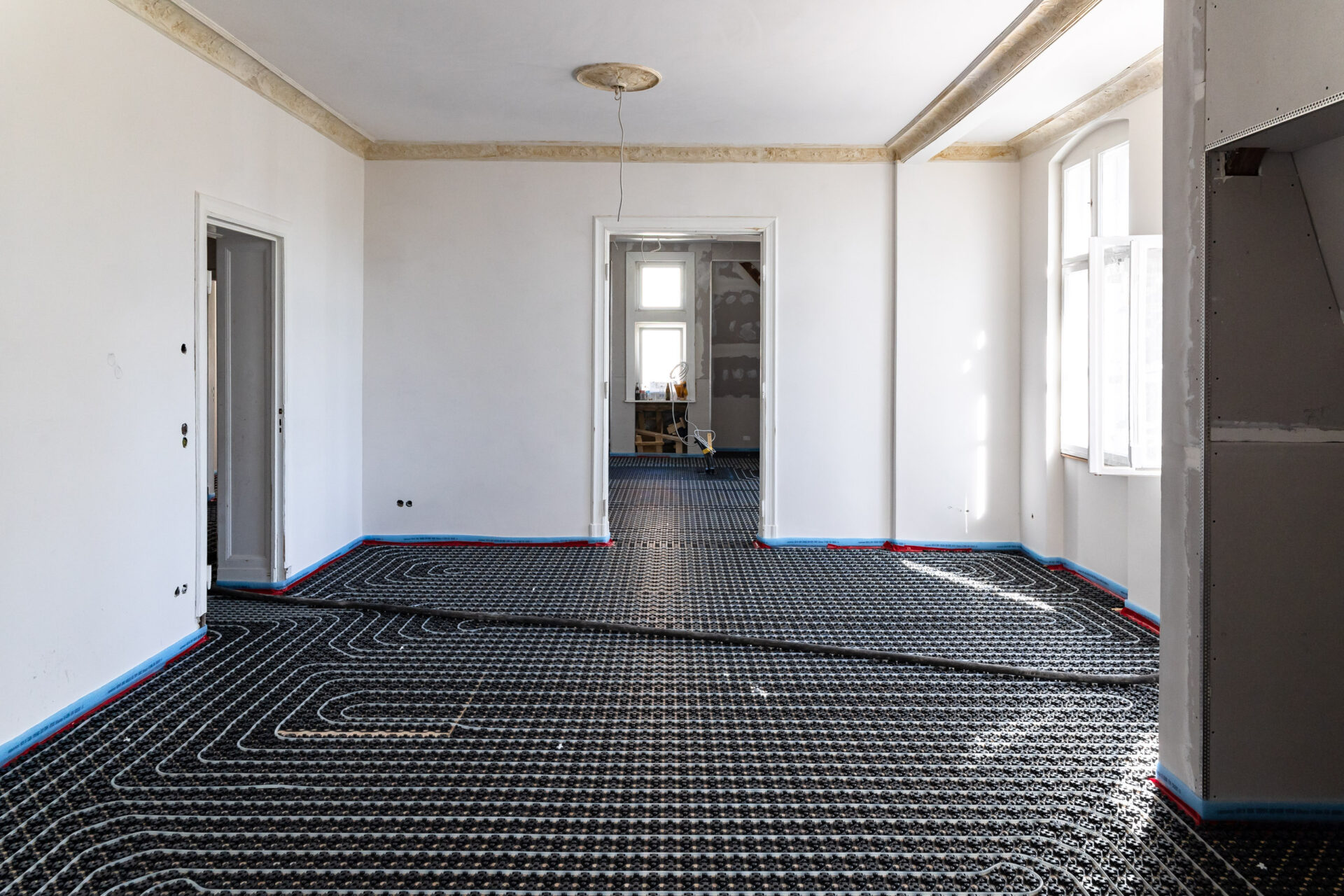 Unmöblierter Raum mit weißen Wänden und sichtbarer Fußbodenheizung, die mit den angrenzenden Räumen verbunden ist; durch die Fenster fällt Sonnenlicht ein.