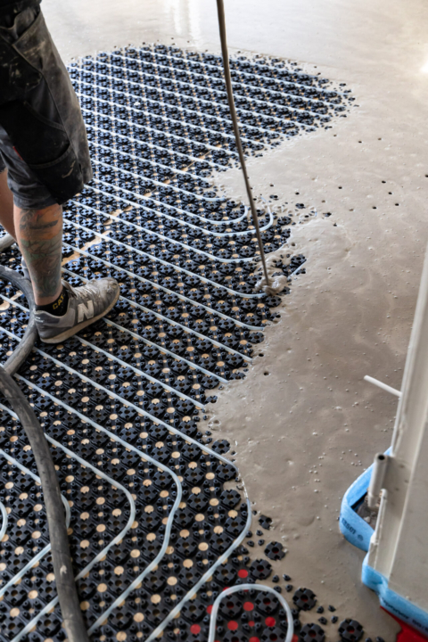 Ein Arbeiter gießt Beton über eine Fußbodenheizung mit freiliegenden Rohren und schwarzen Kunststoffgitterplatten.