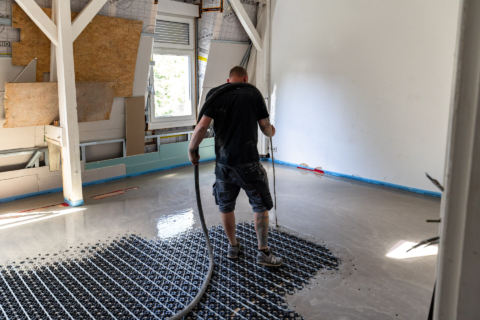 Ein Arbeiter gießt und verteilt mit einem Schlauch selbstnivellierenden Beton auf einem Boden mit Fußbodenheizung in einem teilweise renovierten Raum.