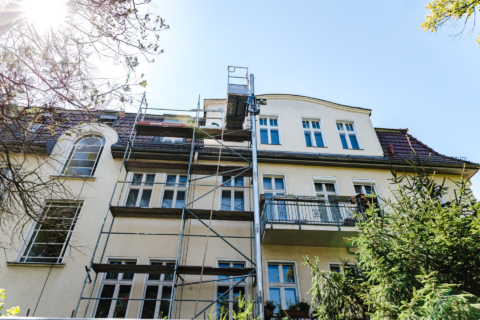 Ein Gerüst ist an der Außenseite eines mehrstöckigen Wohngebäudes unter blauem Himmel aufgebaut, wobei die Bäume im Vordergrund teilweise sichtbar sind.