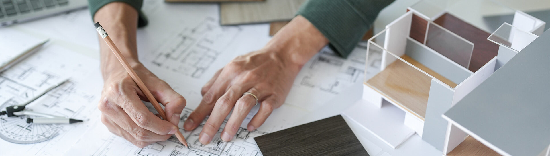 Hände zeichnen auf einem Architekturplan mit Holzmustern und Modellhaus im Hintergrund – individuelle Raumplanung.
