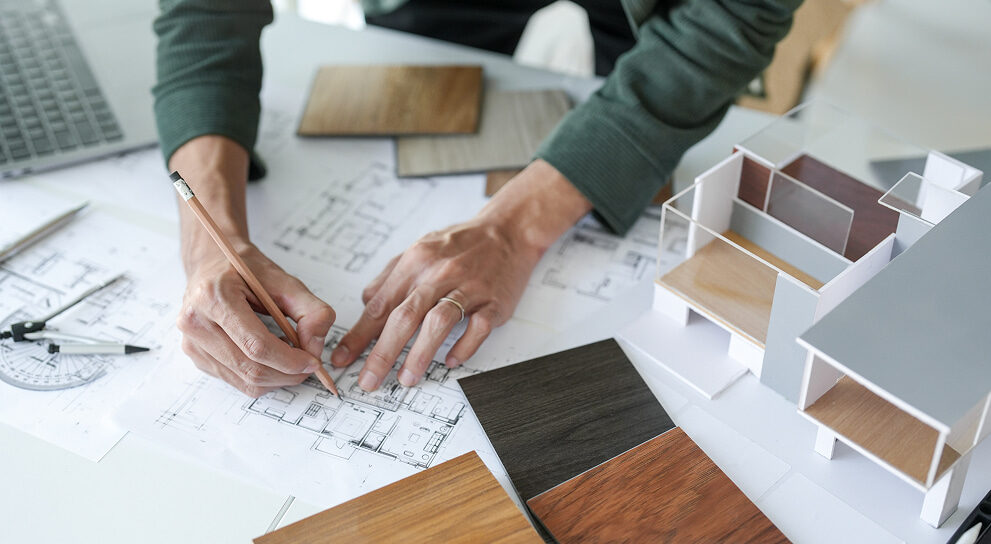 Hände zeichnen auf einem Architekturplan mit Holzmustern und Modellhaus im Hintergrund – individuelle Raumplanung.