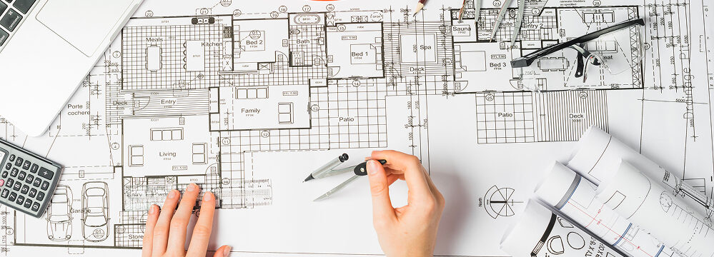 Architektenhände beim Zeichnen eines Bauplans mit Zirkel, Lineal und Grundriss auf dem Tisch.