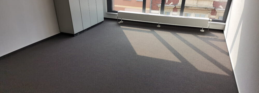 Büro mit frisch verlegtem dunklem Teppichboden, großen Fenstern und modernem Heizkörper unter dem Fenster.