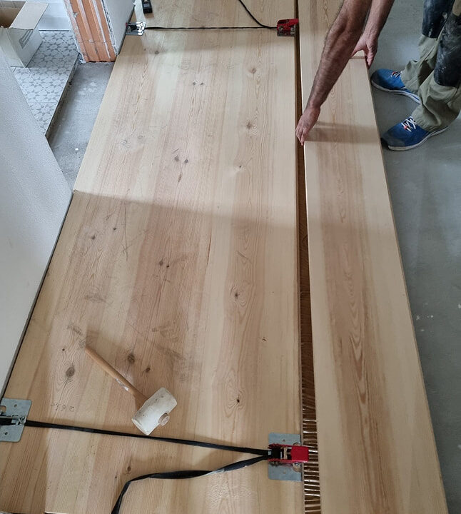 Handwerker beim Verlegen eines Dielenbodens mit Spannwerkzeug und Gummihammer auf einer Baustelle.