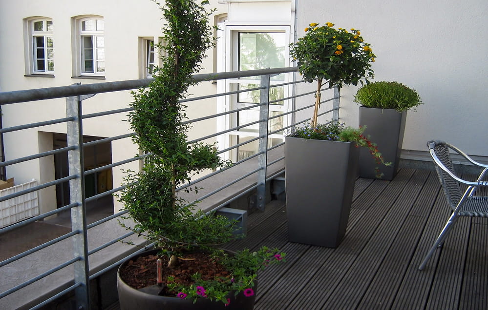 Ein kleiner Balkon mit Metallgeländer, moderne graue Pflanzkübel mit üppigen Grünpflanzen und schlichte Metallstühle auf warmen Holzdielen.