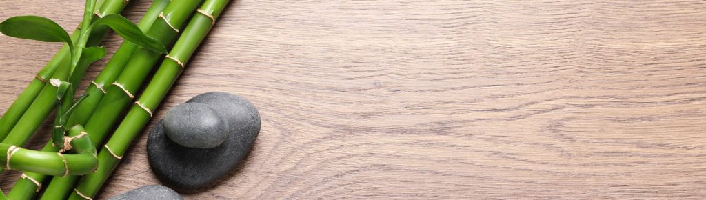Drei grüne Bambushalme und zwei glatte schwarze Steine werden auf einer hellen Holzfläche platziert, mit viel freiem Platz auf der rechten Seite.