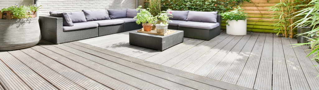 Terrasse im Freien mit geflochtenem Sektionssofa, grauen Kissen, Topfpflanzen auf einem Tisch und einer Holzterrasse, umgeben von Grünpflanzen und einem Holzzaun.