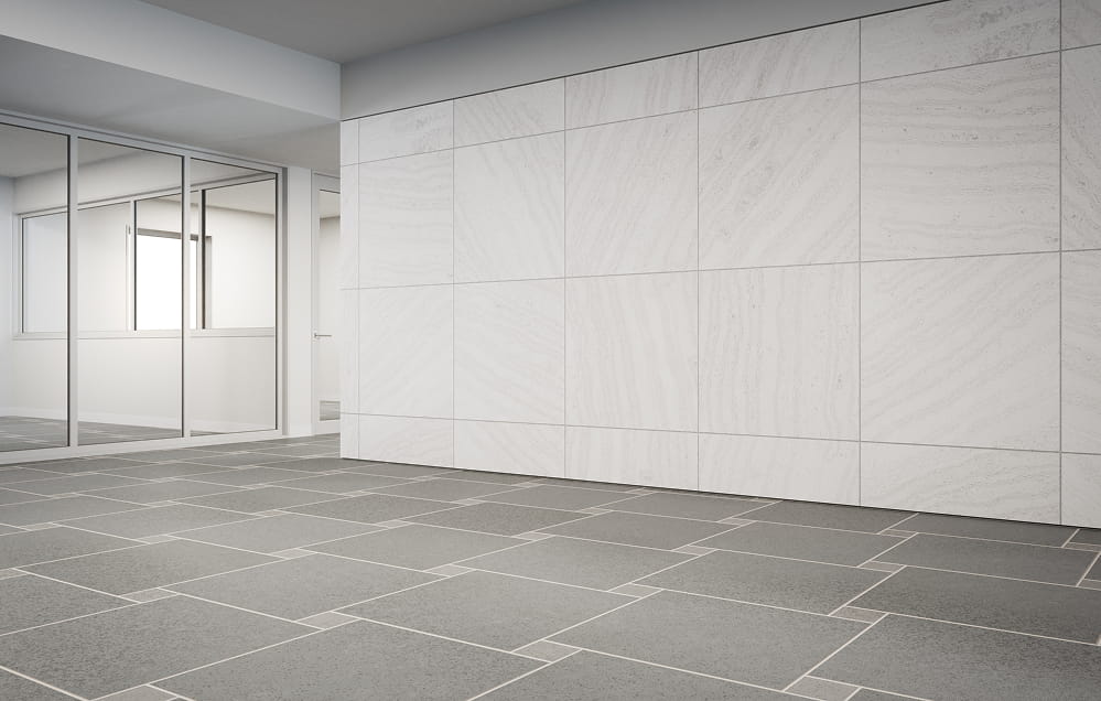 Leerer, moderner Innenraum mit großer weiß gefliester Wand, grauem Fliesenboden und Glastrennwand, die einen weiteren Raum freigibt.