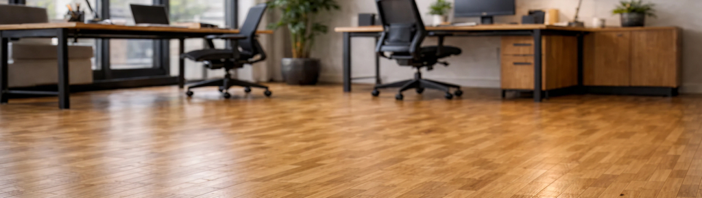 Moderne Büroräume mit Industrieparkett-Holzboden, zwei Schreibtischen mit Bürostühlen, Computern, Lampen, Pflanzen und großen Fenstern, die für Tageslicht sorgen.