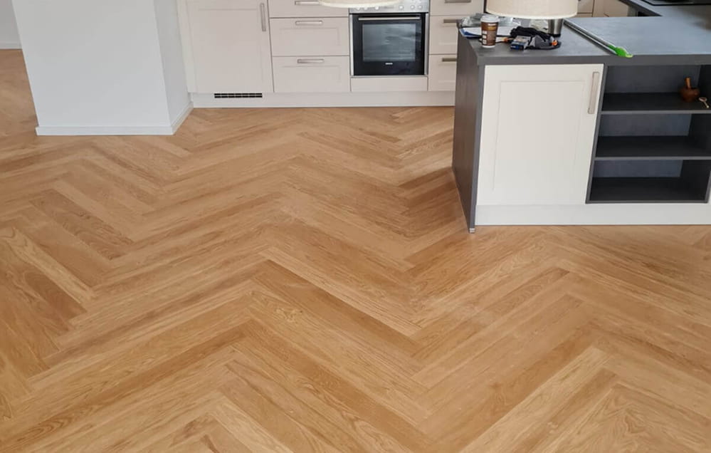 Eine Küche mit weißen Schränken und einer Insel, die mit einem hellen Parkettboden mit Fischgrätenmuster ausgestattet ist.