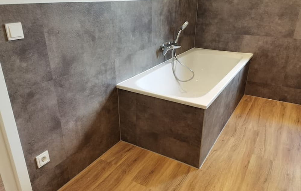 Eine weiße Badewanne mit einer Handbrause steht vor gefliesten Wänden in einem Badezimmer mit Vinylboden in Holzoptik.