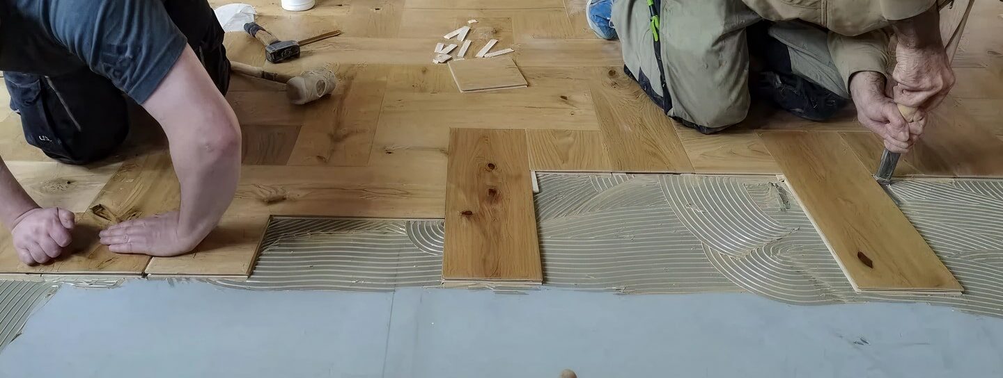 Zwei Personen verlegen Holzdielen, indem sie Klebstoff auf den Unterboden auftragen und die Dielen vorsichtig anbringen.