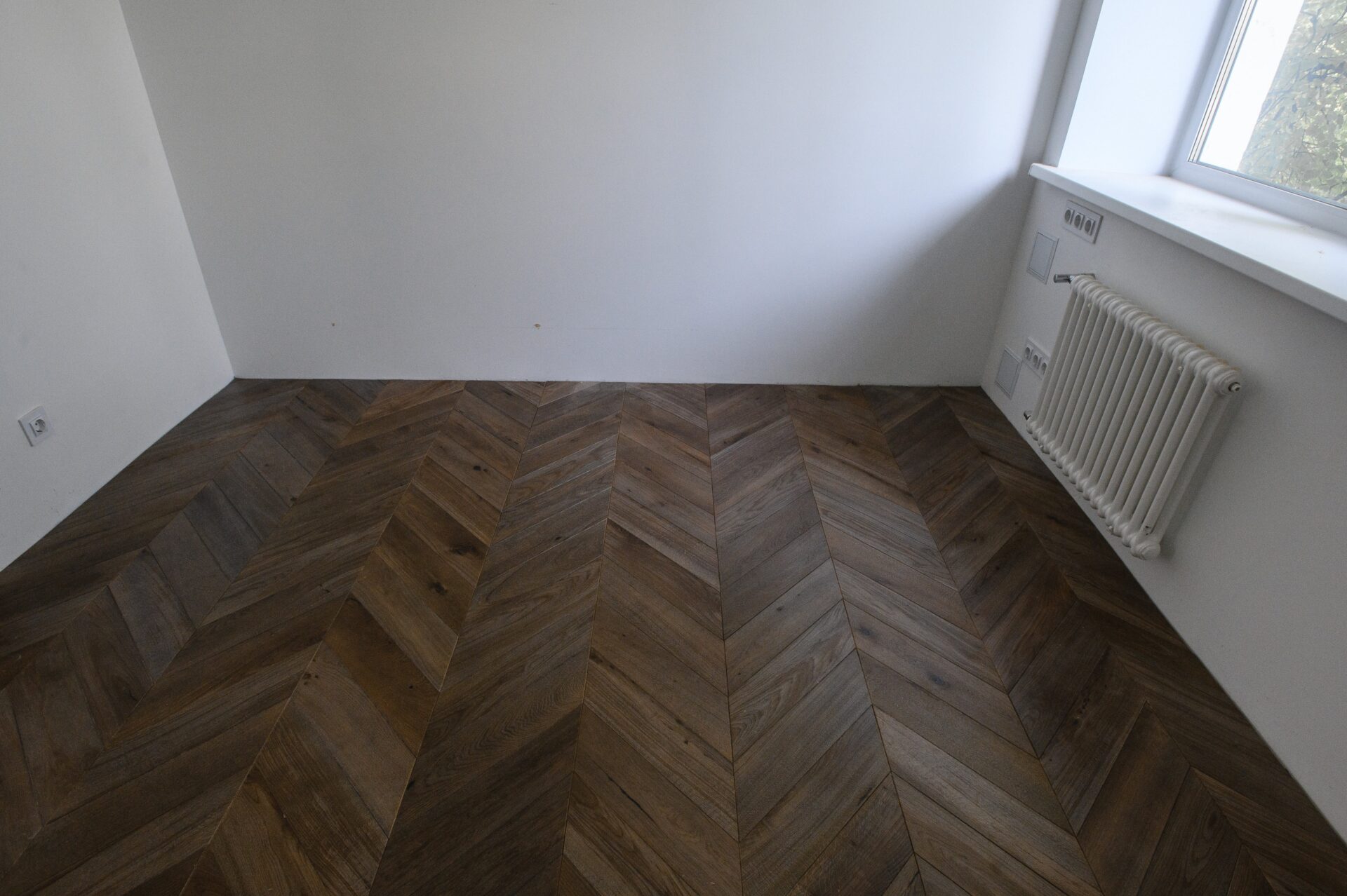 Ein leerer Raum mit weißen Wänden, einem Parkettfußboden, einem Heizkörper unter dem Fenster und sichtbaren Steckdosen.