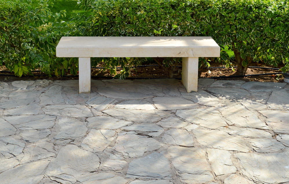 Eine Steinbank steht auf einer Natursteinterrasse vor grünen Büschen auf einer gepflasterten Fläche in einem Außenbereich.