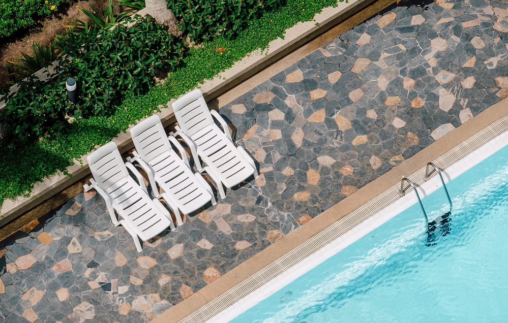 Drei weiße Plastiksessel stehen in einer Reihe neben einem Swimmingpool mit gemusterten Steinfliesen, die die Poolumrandung bilden, und üppigen Grünpflanzen in der Nähe.