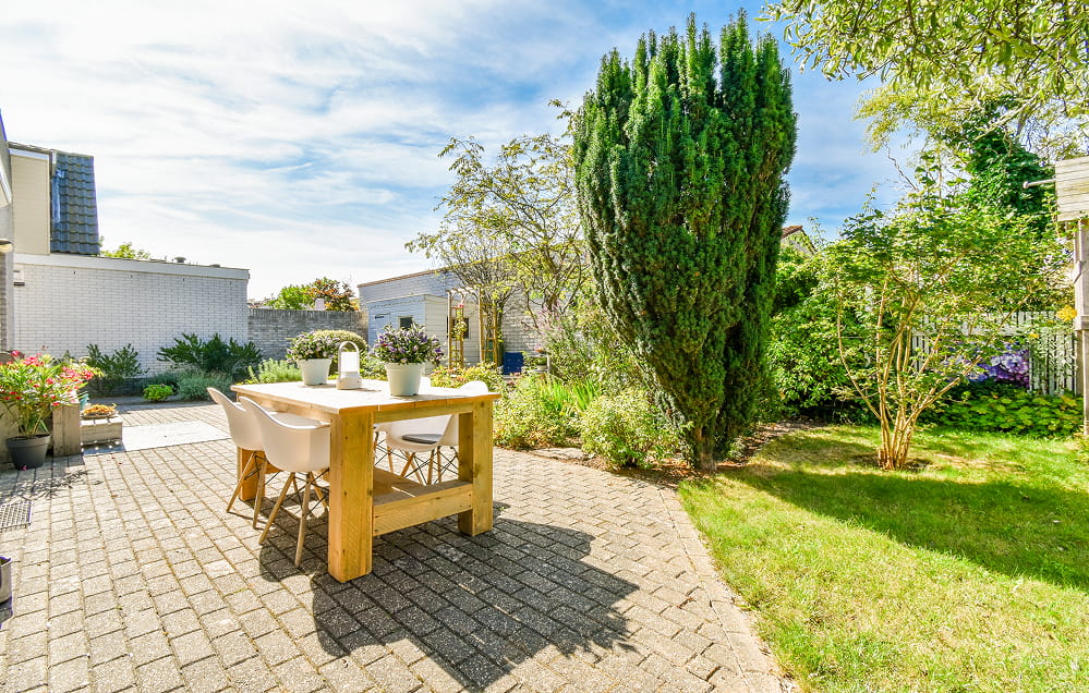 Ein Holztisch mit weißen Stühlen steht auf einer sonnigen Terrasse mit Pflaster, umgeben von grünem Gras, hohen Bäumen und üppigen Gartenpflanzen.