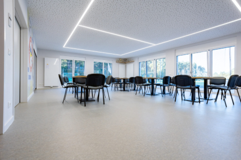 Ein moderner, geräumiger Raum mit mehreren runden Tischen und schwarzen Stühlen, großen Fenstern, die natürliches Licht hereinlassen, und geometrischen LED-Deckenleuchten.