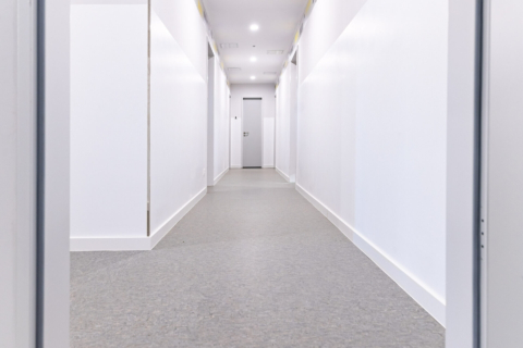 Ein sauberer, hell erleuchteter Flur mit weißen Wänden, grauem Bodenbelag und geschlossenen Türen auf beiden Seiten, die zu einer geschlossenen Tür am Ende führen.