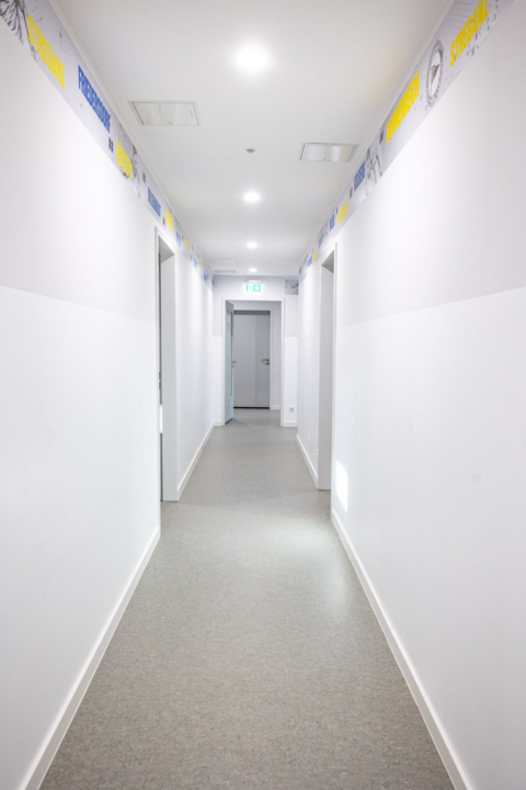 Ein langer, weißer Flur mit geschlossenen Türen auf beiden Seiten, Neonröhren über dem Kopf und grauem Boden. Schilder mit blauem und gelbem Text säumen den oberen Teil der Wände.