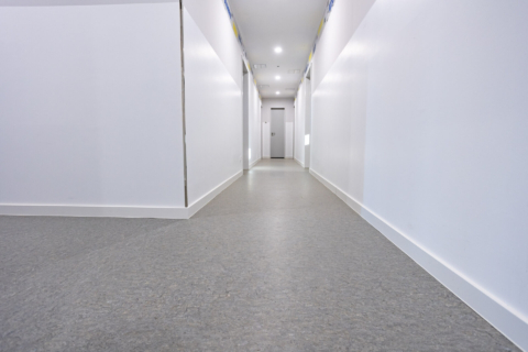 Ein sauberer, leerer Flur mit weißen Wänden, einem grauen Fußboden und einer geschlossenen Tür am anderen Ende. Helle Deckenleuchten erhellen den Raum.