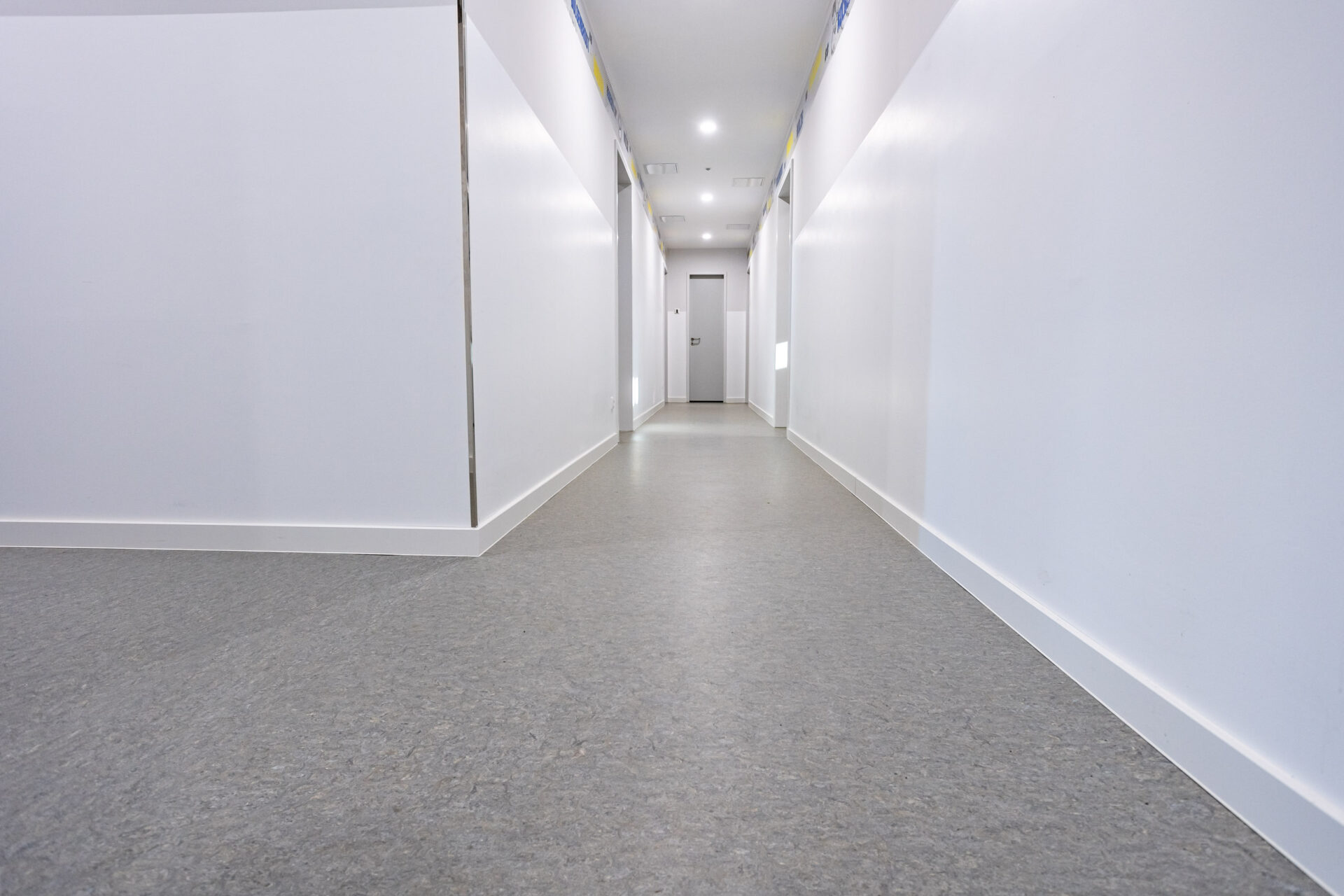 Ein sauberer, leerer Flur mit weißen Wänden, einem grauen Fußboden und einer geschlossenen Tür am anderen Ende. Helle Deckenleuchten erhellen den Raum.