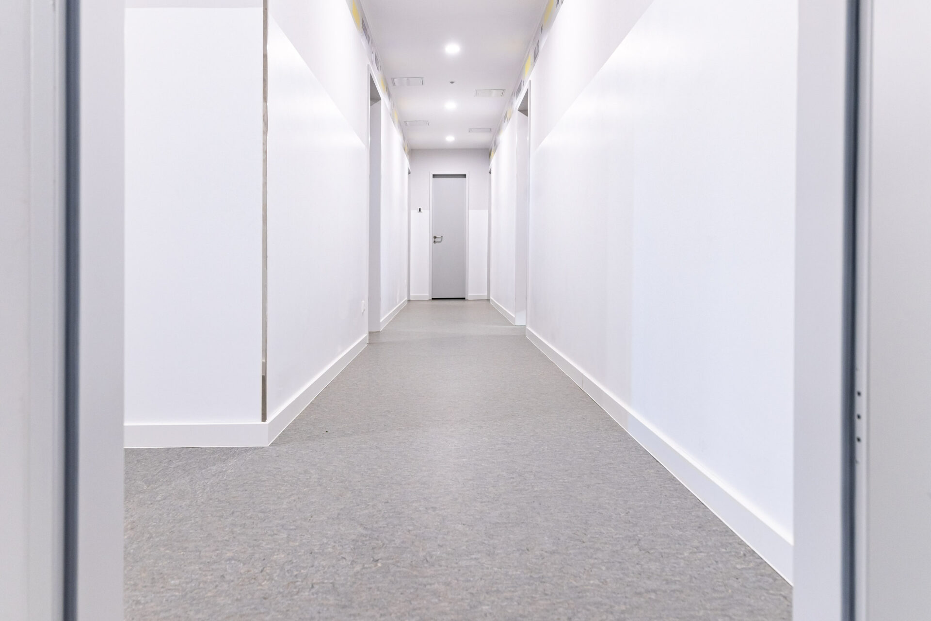 Ein sauberer, hell erleuchteter Flur mit weißen Wänden, grauem Bodenbelag und geschlossenen Türen auf beiden Seiten, die zu einer geschlossenen Tür am Ende führen.