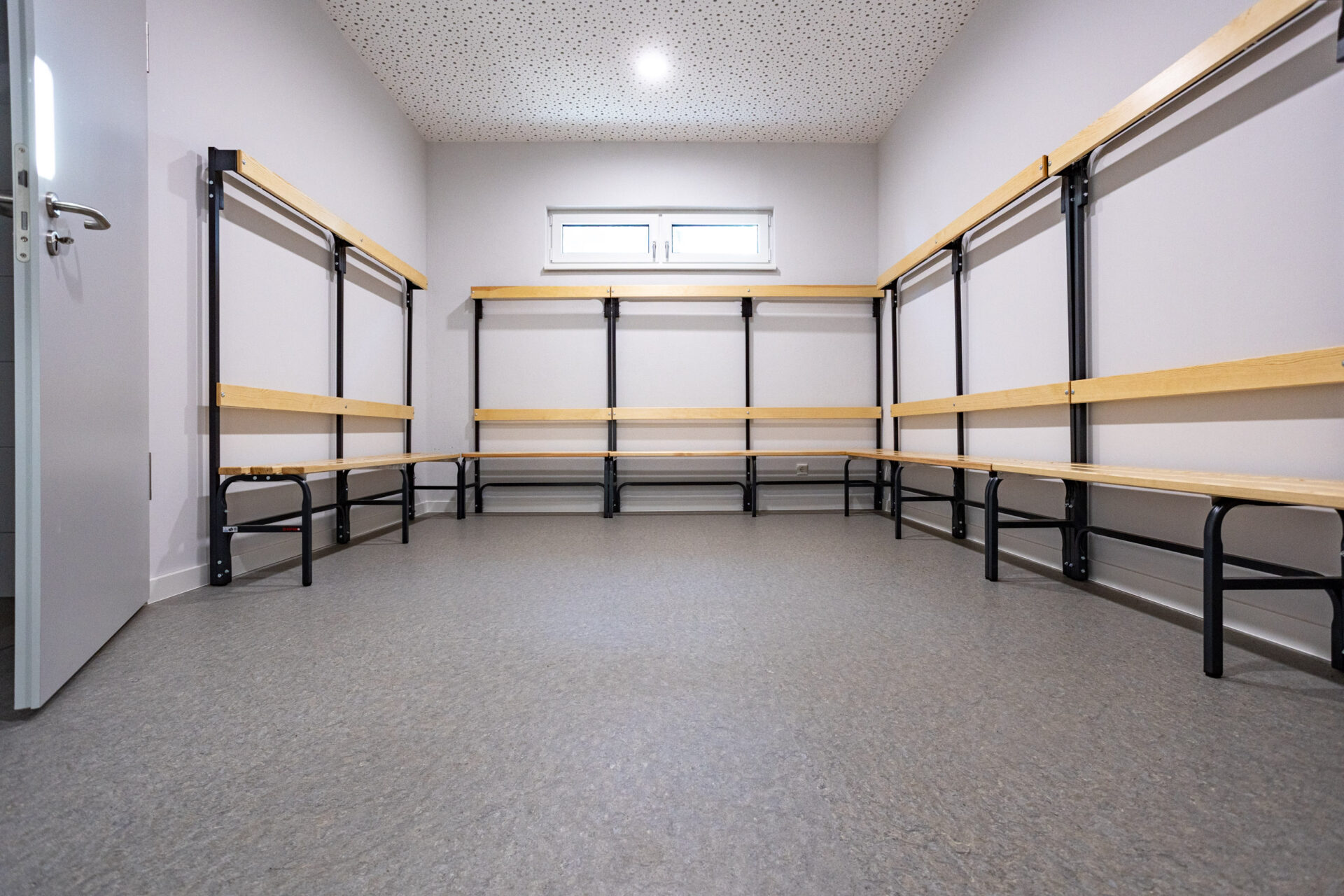 Leere Umkleidekabine mit hellen Wänden, Bänken an drei Seiten und einem kleinen Fenster darüber; auf der linken Seite ist eine offene Tür zu sehen.