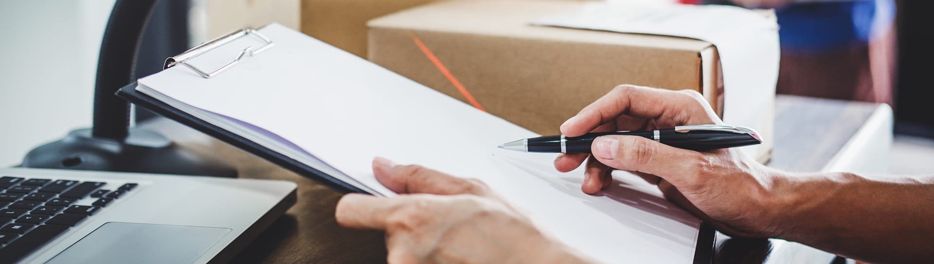 Eine Person mit einem Stift schreibt auf einem Klemmbrett neben einem Laptop und einer Versandbox auf einem Schreibtisch.