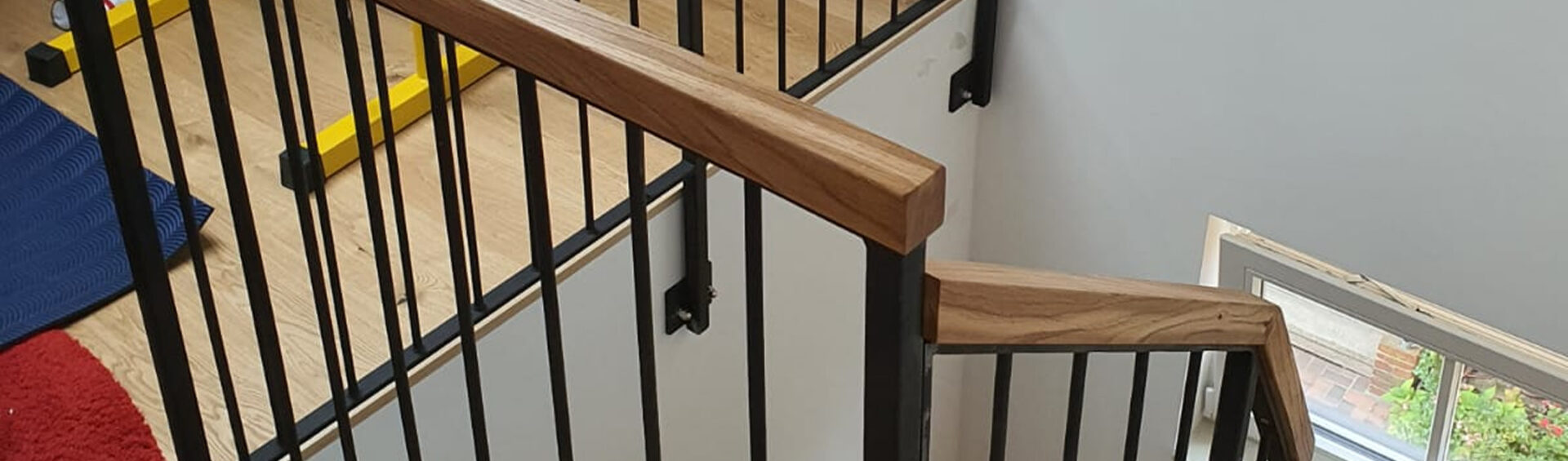 Treppenhandlauf aus massivem Eichenholz in Kombination mit einem schwarzen Stahlgeländer, montiert in einem modernen Treppenhaus mit Fenster und Holzboden.
