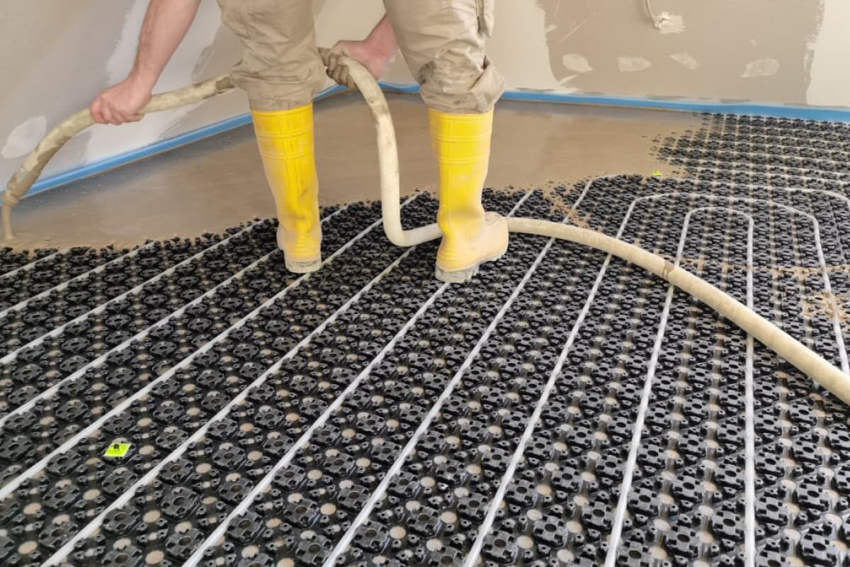 Ein Handwerker mit gelben Gummistiefeln trägt Fließestrich über einer installierten Fußbodenheizung mit Noppenplatten auf. Die Estrichmasse wird mithilfe eines Schlauchs gleichmäßig verteilt.