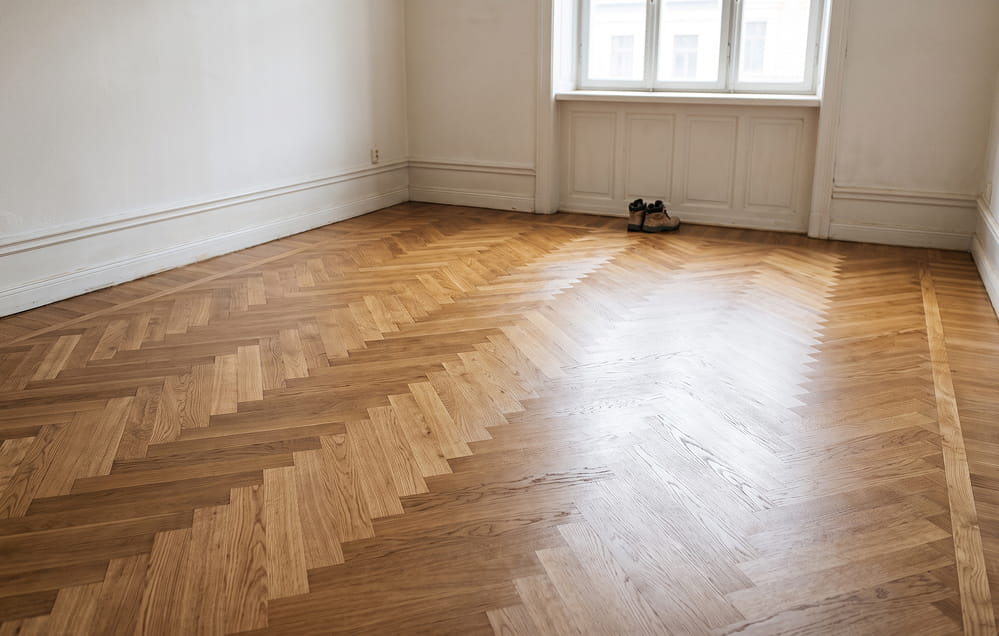 Ein leerer Raum mit frisch versiegeltem Fischgrätenparkett, weißen Wänden, einem Fenster, durch das Tageslicht hereinfällt, und einem einzelnen Paar Schuhe neben dem Fenster.