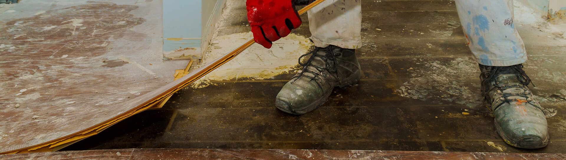 Handwerker mit roten Handschuhen entfernt alten Bodenbelag in einem renovierungsbedürftigen Raum.