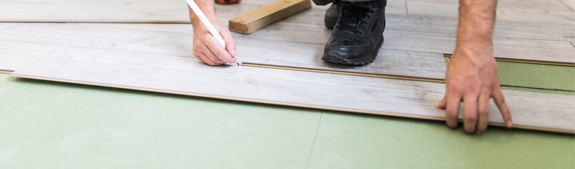 Handwerker markiert Laminatdiele mit Bleistift zum Zuschneiden, daneben Holzklotz und Werkzeug auf grüner Trittschalldämmung.