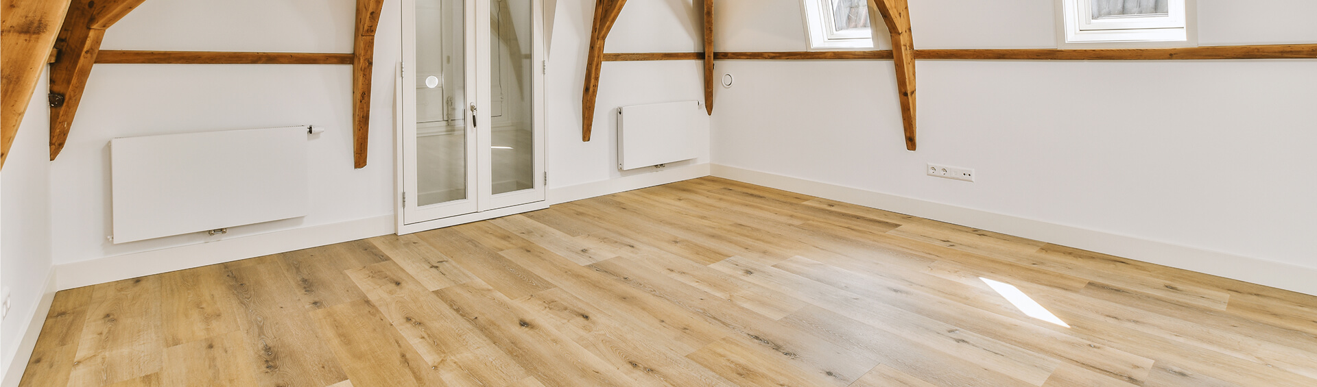 Leerer Dachgeschossraum mit hellem Holzboden, freiliegenden Holzbalken und weißer Doppeltür, Dachfenster lassen Licht herein.