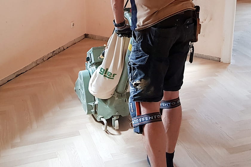 Eine Person, die kurze Hosen und Kniestützen trägt, bedient eine Schleifmaschine auf einem hellen Holzboden in einem Raum.