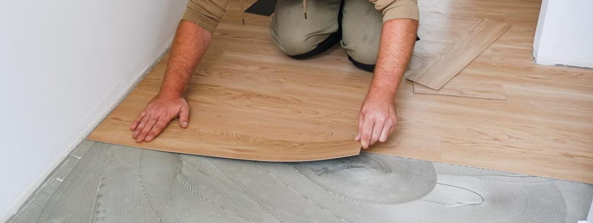 Handwerker verklebt eine Parkettdiele am Randbereich einer Wand mit sichtbarem Kleber auf dem Untergrund.