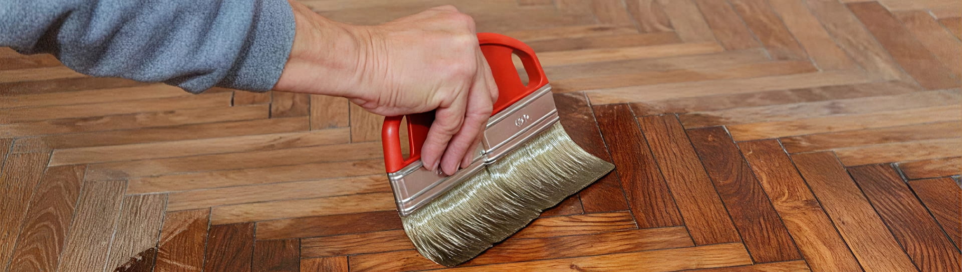 Ein Mann der mit einem breitem Pinsel ein geschliffenes Parkett mit Lack versiegelt.