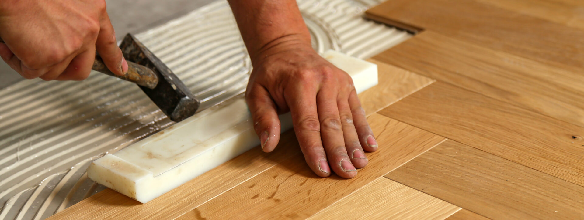 Nahaufnahme eines Handwerkers beim präzisen Verlegen von Parkett mit Kleber und Hammer.