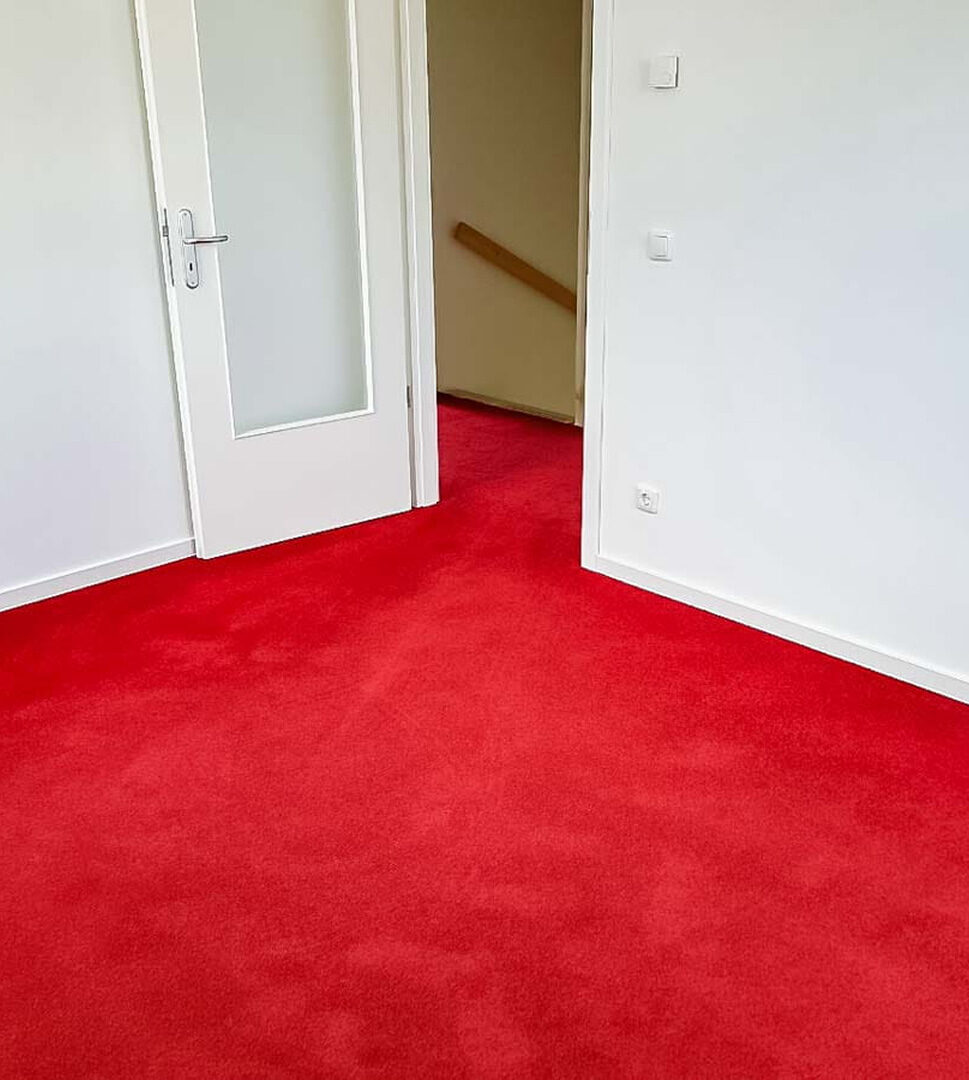 Roter Teppichboden in einem hellen Raum mit weißer Tür und Blick in einen weiteren Raum mit gleichem Teppich.