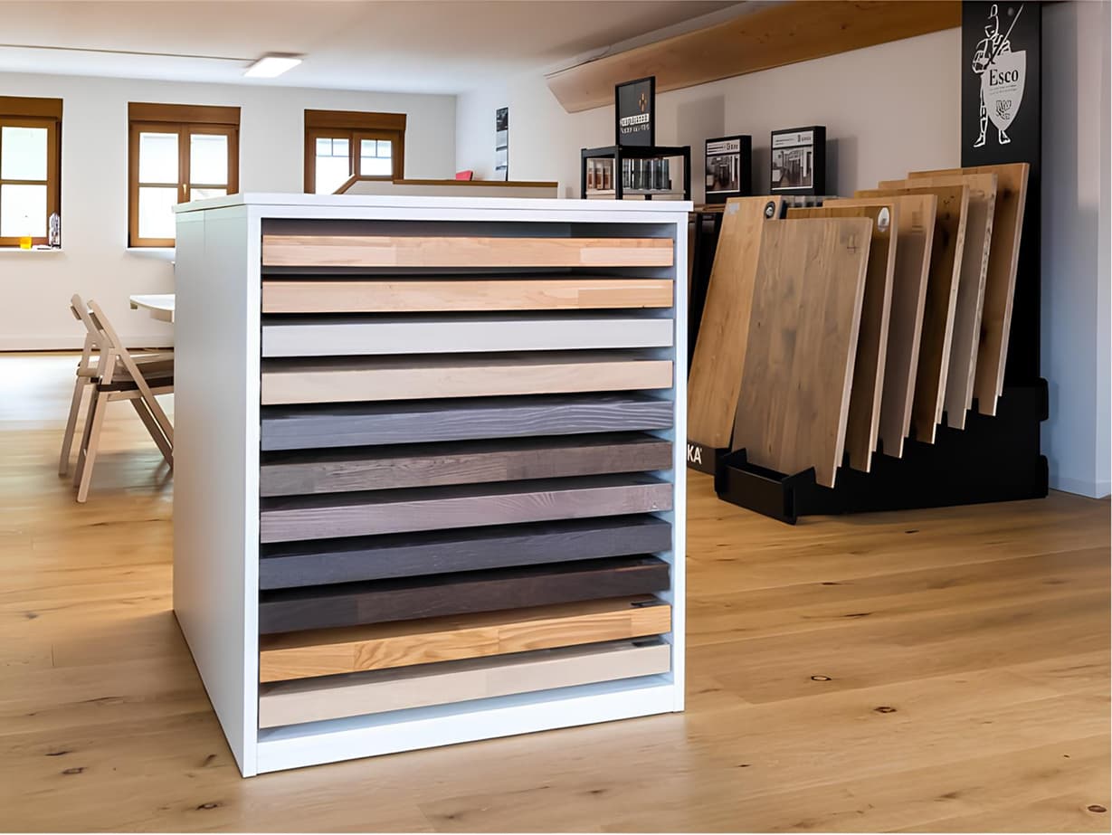 Moderner Showroom der Domke Parkett GmbH in Berlin mit einer stilvollen Ausstellung verschiedener Parkettmuster. Im Vordergrund steht ein Musterblock mit Holzproben in unterschiedlichen Farben und Oberflächenstrukturen. Im Hintergrund sind große Holztafeln mit Parkettmustern sowie ein gemütlicher Besprechungsbereich mit Holzboden zu sehen.