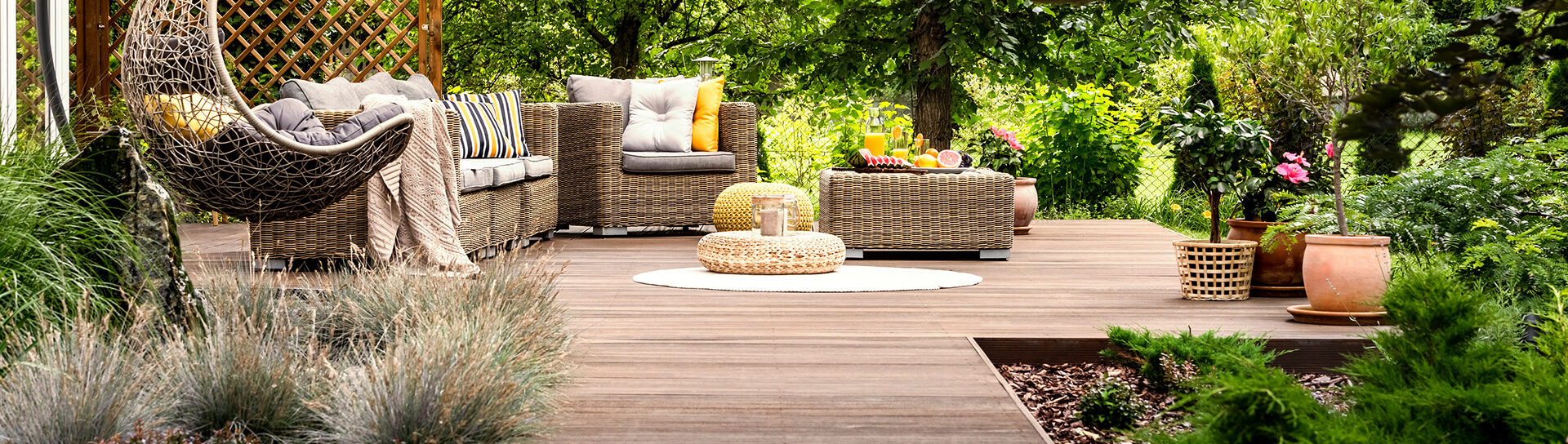 Gemütliche Gartenterrasse mit Holzbelag, Rattanmöbeln und Hängesessel inmitten grüner Pflanzen.