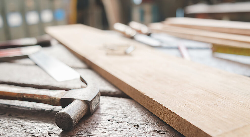 Nahaufnahme einer Holzplatte mit Hammer und Säge auf einer Werkbank – Symbolbild für präzise Tischlerarbeiten und handwerkliches Arbeiten mit Holz.