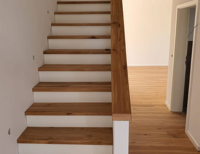 Eine Holztreppe mit weißen Setzstufen und einem passenden Handlauf aus Holz führt neben einem hellen Hartholzboden in einem minimalistisch gestalteten Innenraum nach oben.