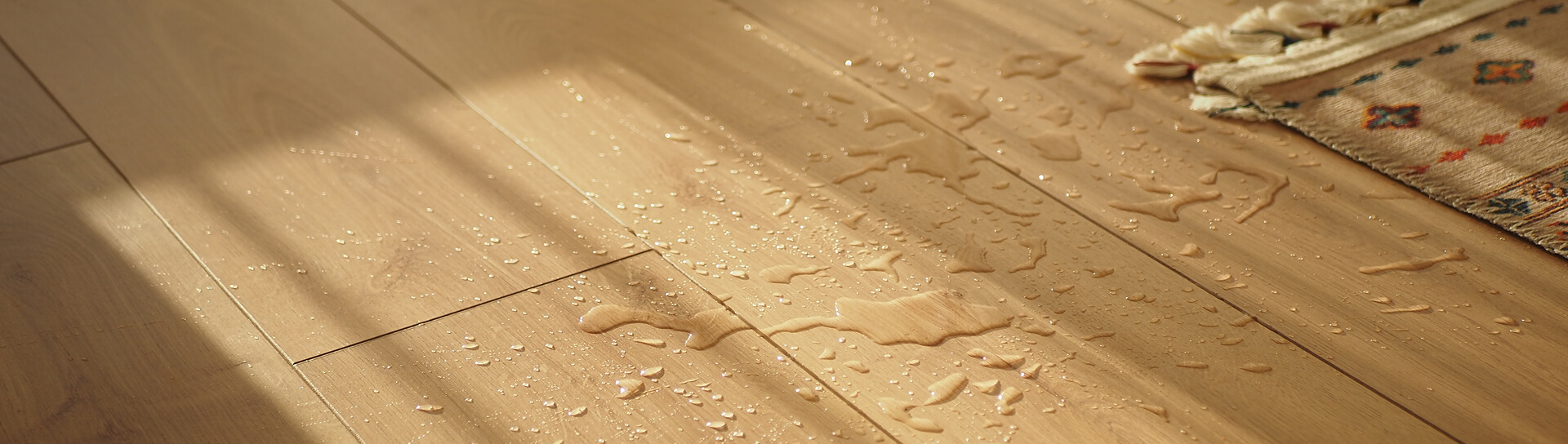 Wassertropfen auf hellem Holzparkett, die im Sonnenlicht glänzen, rechts im Bild die Ecke eines gemusterten Teppichs.